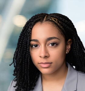 A young woman with braided hair and a neutral expression, wearing a light gray blazer, poses in front of a blurred modern office background with soft lighting.