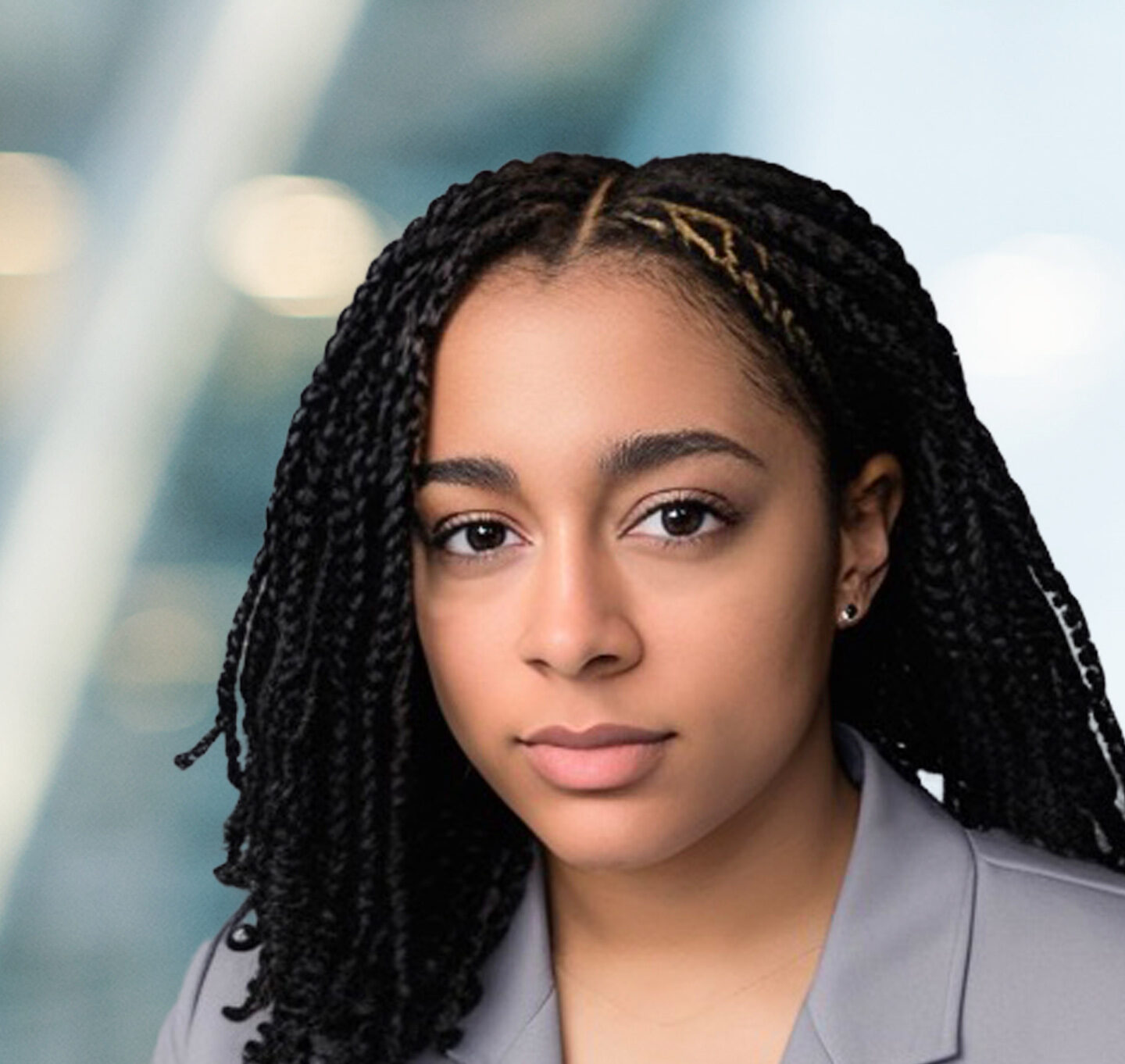 A young woman with braided hair and a neutral expression, wearing a light gray blazer, poses in front of a blurred modern office background with soft lighting.