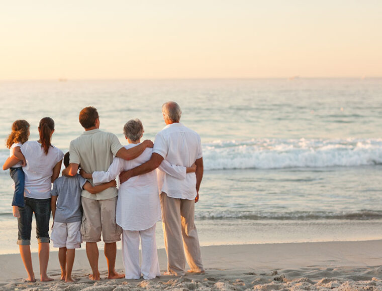 A family of six, including adults, seniors, and children, stands on the beach facing the ocean at sunset, with their arms around each other, enjoying the view together.