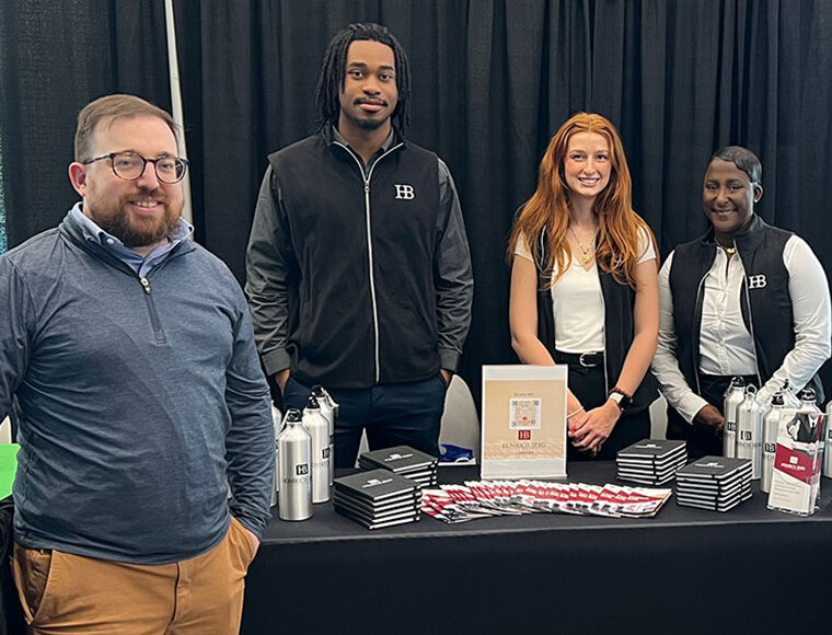 Four people stand behind a table displaying water bottles, notebooks, and brochures at a booth. Three wear black vests with “HB.” They are smiling, with a black curtain and windows in the background.