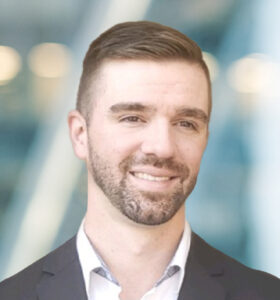 A man with short brown hair and a trimmed beard, wearing a dark blazer and light shirt, smiles slightly while standing in front of a blurred background with blue and white tones.