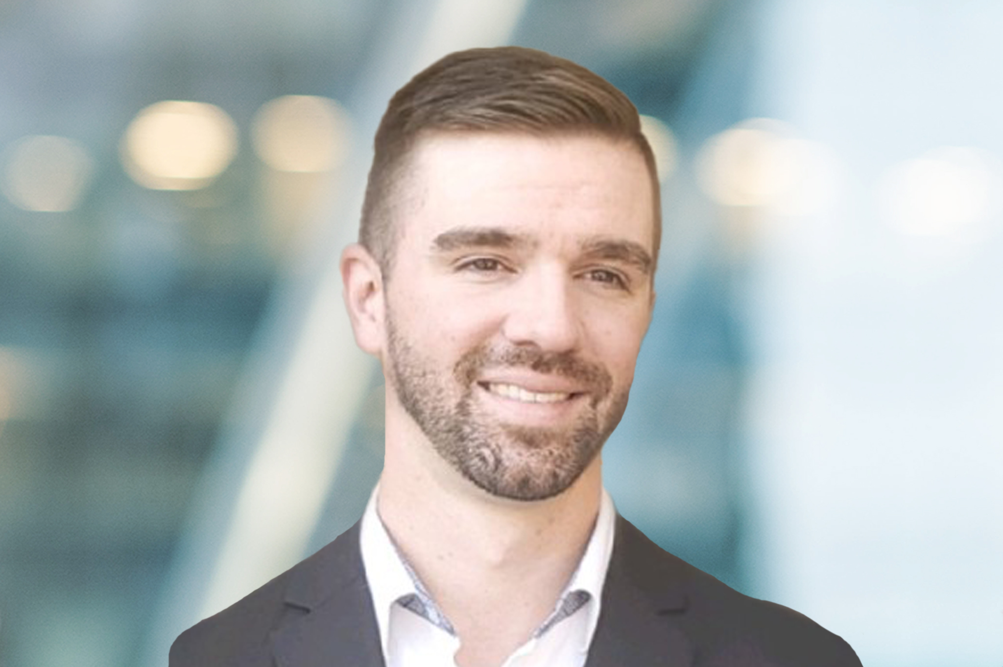 A man with short brown hair and a trimmed beard, wearing a dark blazer and light shirt, smiles slightly while standing in front of a blurred background with blue and white tones.