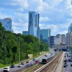 A busy highway with multiple lanes of traffic runs alongside a railway track, leading toward a city skyline with modern high-rise buildings and lush green trees under a partly cloudy sky.