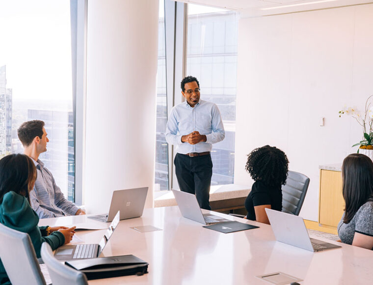 A man stands and speaks to four seated colleagues around a conference table in a modern office with large windows. Laptops are open on the table, and sunlight fills the room.
