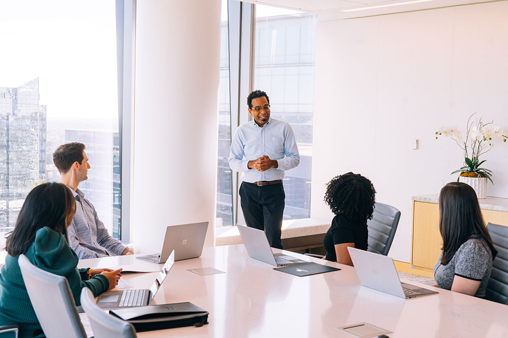 A man stands and speaks to four seated colleagues around a conference table in a modern office with large windows. Laptops are open on the table, and sunlight fills the room.