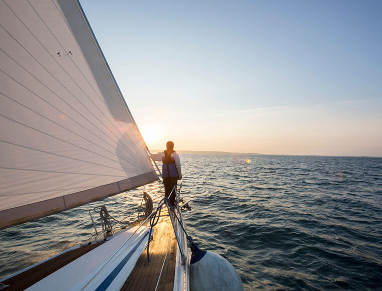 A person stands on the deck of a sailboat holding the mast, looking towards the sunset over calm, open water, with the boats sail and rigging visible in the foreground.