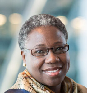 A woman with short gray hair and glasses smiles at the camera, wearing a plaid scarf and earrings, with a blurred background of soft lights and blue tones.