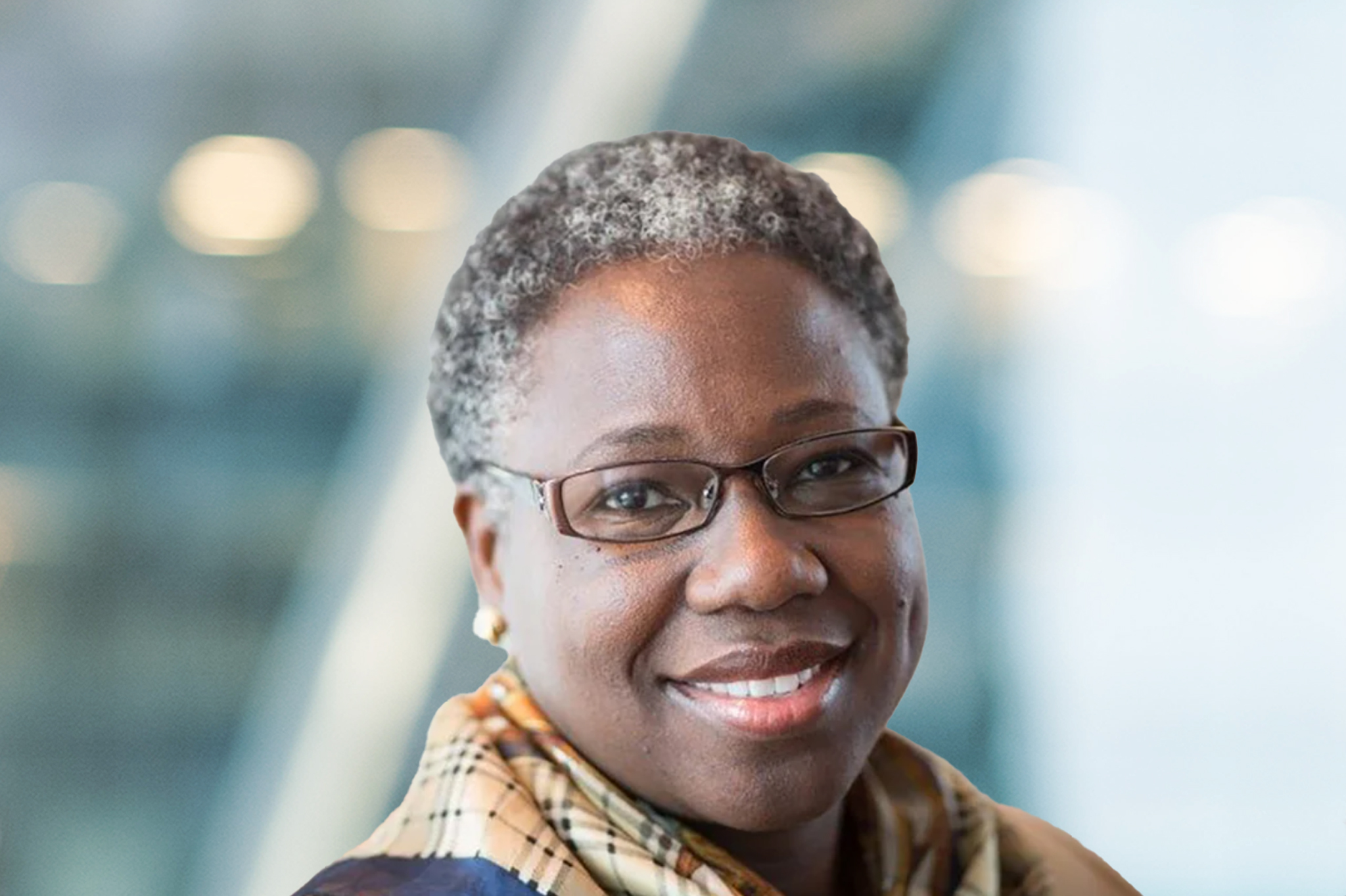 A woman with short gray hair and glasses smiles at the camera, wearing a plaid scarf and earrings, with a blurred background of soft lights and blue tones.