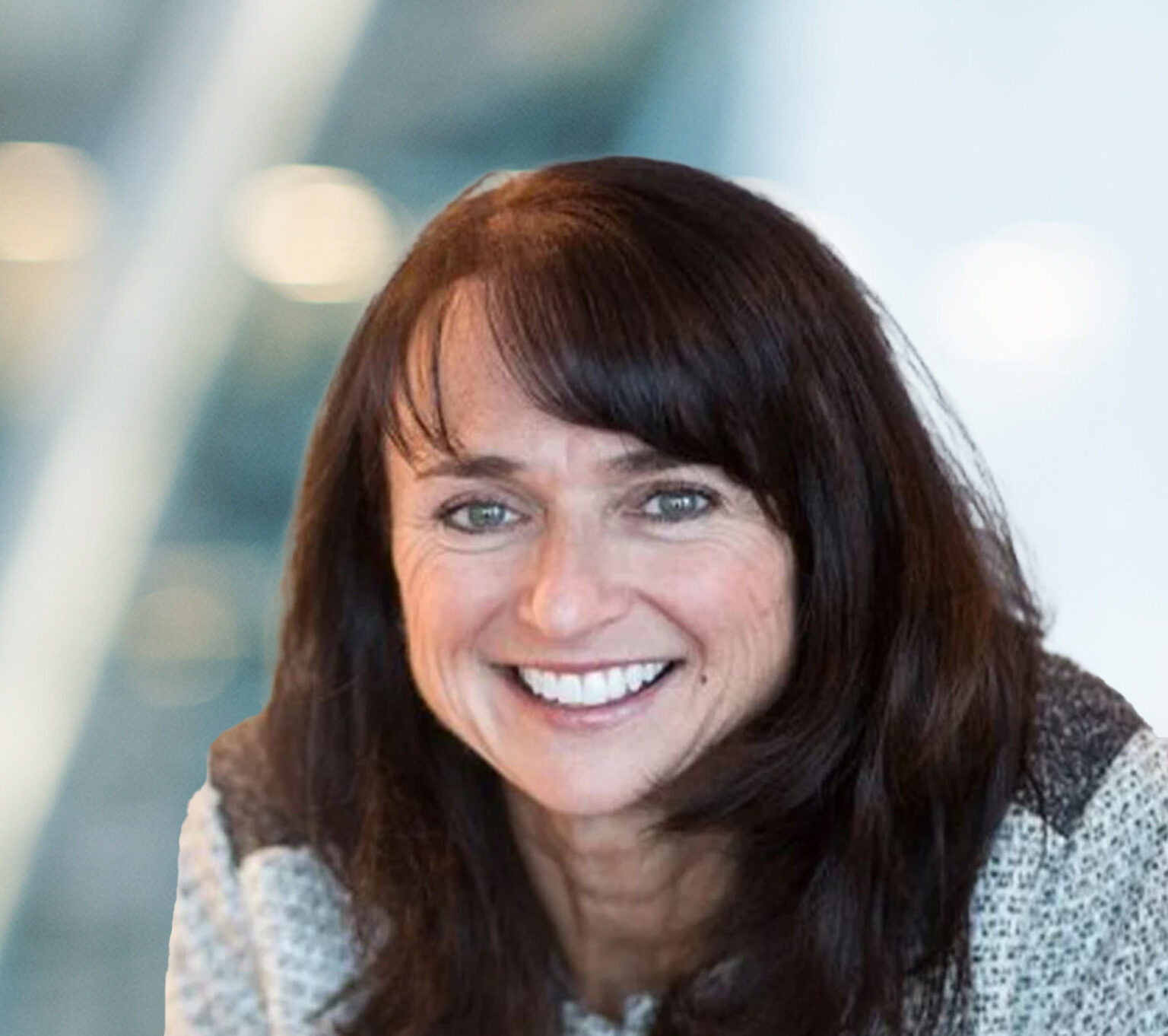 A woman with long brown hair and bangs is smiling, wearing a gray sweater. The background is blurred with soft lights and abstract shapes.
