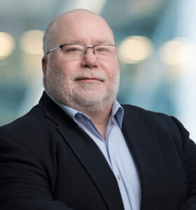 A middle-aged man with a bald head, glasses, and a gray beard stands confidently in a dark suit and light shirt. The background is blurred with soft lights, suggesting a professional setting.