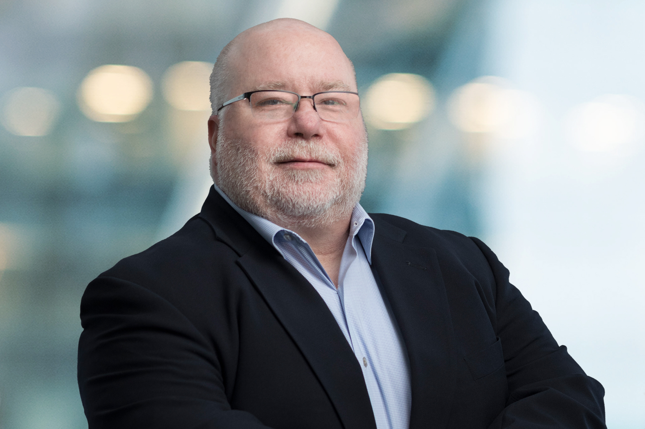 A middle-aged man with a bald head, glasses, and a gray beard stands confidently in a dark suit and light shirt. The background is blurred with soft lights, suggesting a professional setting.