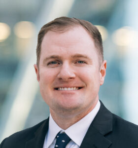 A man in a suit and tie smiling at the camera, standing in front of a blurred modern glass building background.