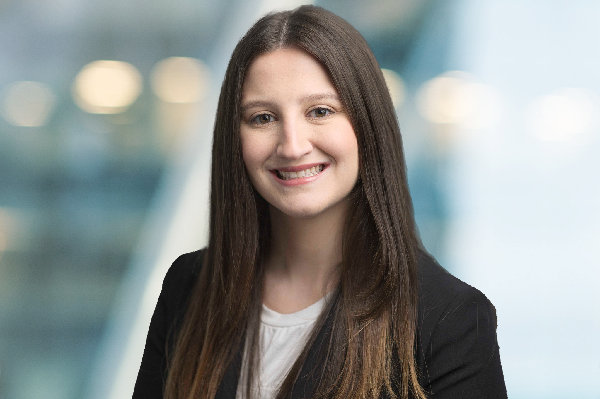 A woman with long straight brown hair, wearing a black blazer over a white top, smiles at the camera against a blurred blue and white background.