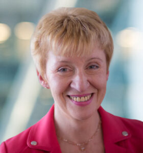 A woman with short, light brown hair smiles at the camera. She is wearing a bright pink blazer and a delicate necklace. The background is softly blurred with blue and white tones.