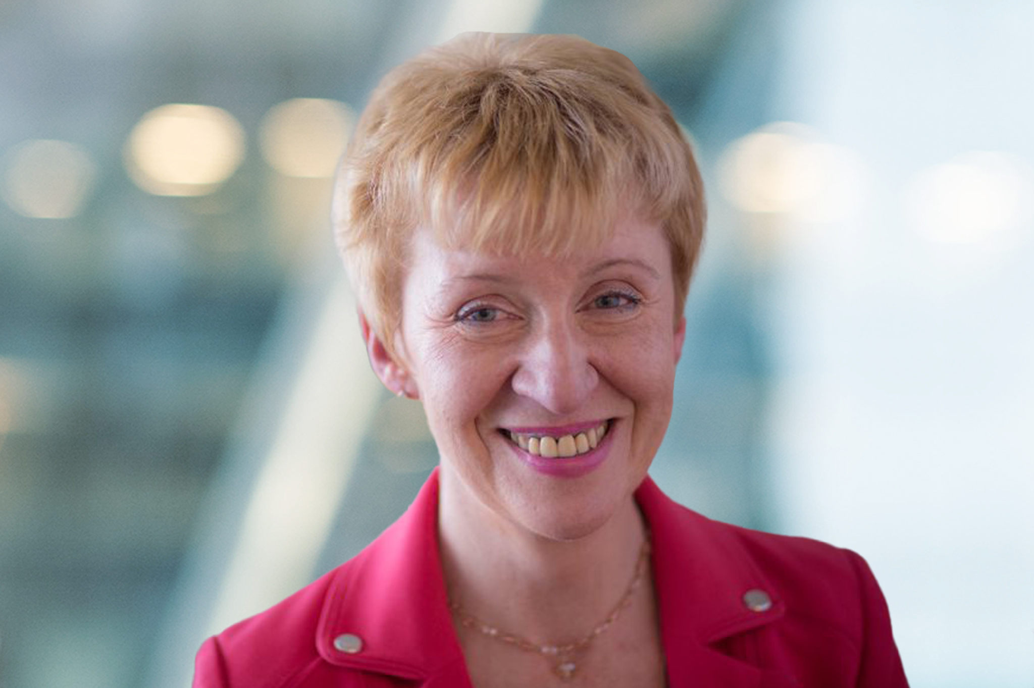 A woman with short, light brown hair smiles at the camera. She is wearing a bright pink blazer and a delicate necklace. The background is softly blurred with blue and white tones.