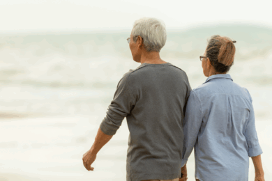 An elderly couple, seen from behind, walks hand in hand along a sandy beach with gentle waves in the background on a cloudy day.