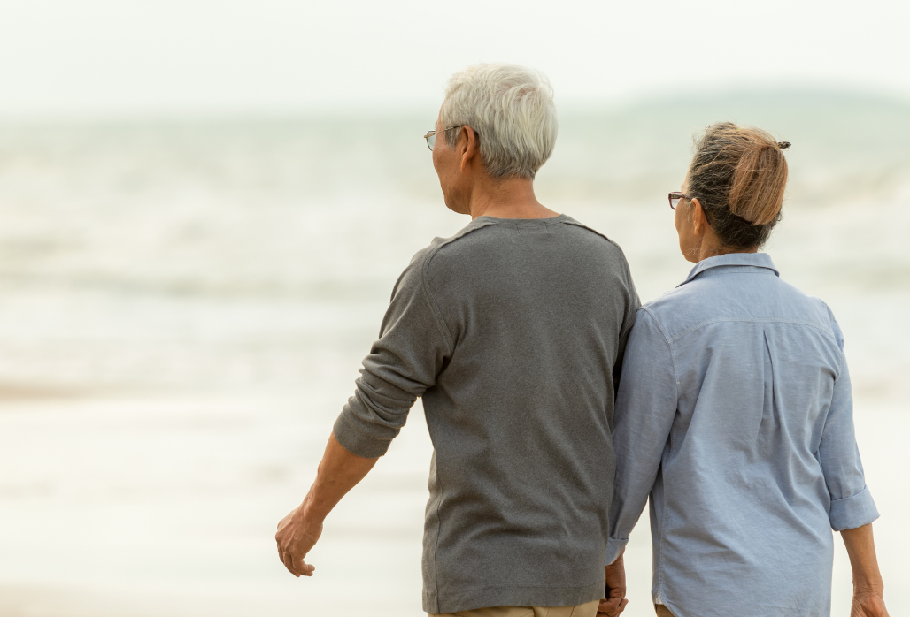 An elderly couple, seen from behind, walks hand in hand along a sandy beach with gentle waves in the background on a cloudy day.