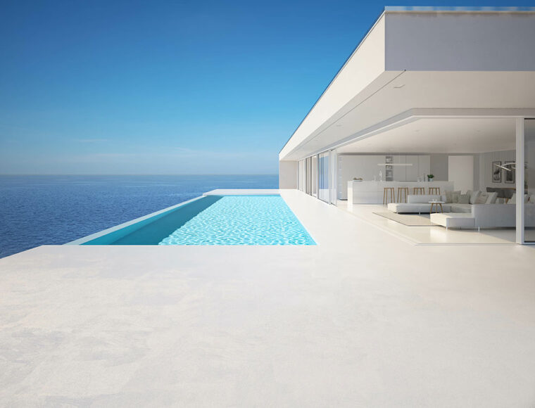 Modern white minimalist house with an infinity pool overlooking a calm blue ocean, featuring floor-to-ceiling glass windows and a spacious open living area. The sky is clear and bright.