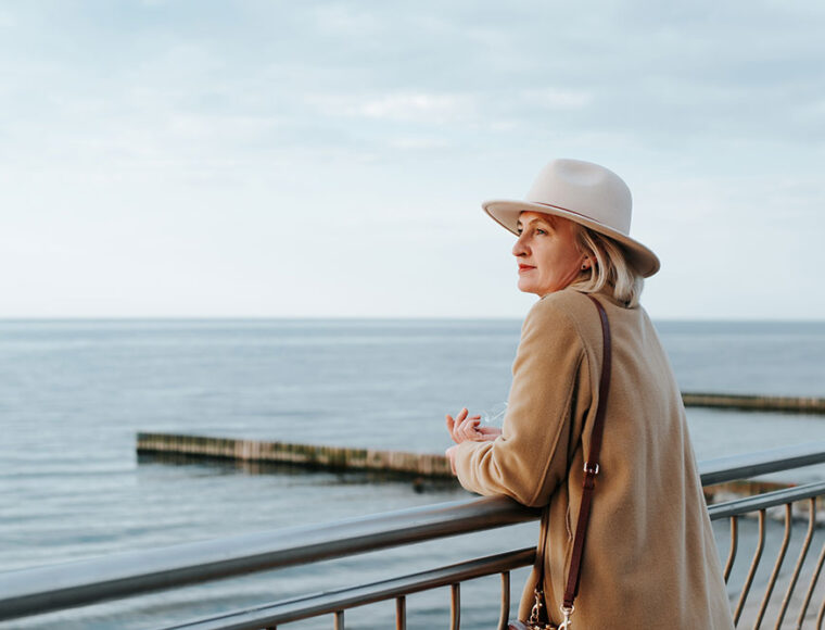 A woman wearing a beige coat and hat leans on a metal railing, gazing thoughtfully at the calm sea under a cloudy sky.