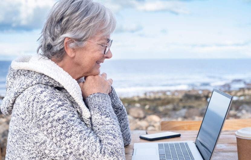 Older woman with gray hair and glasses, wearing a cozy sweater, sits outdoors by the sea, smiling at a laptop on a wooden table. A smartphone and coffee cup are also on the table.