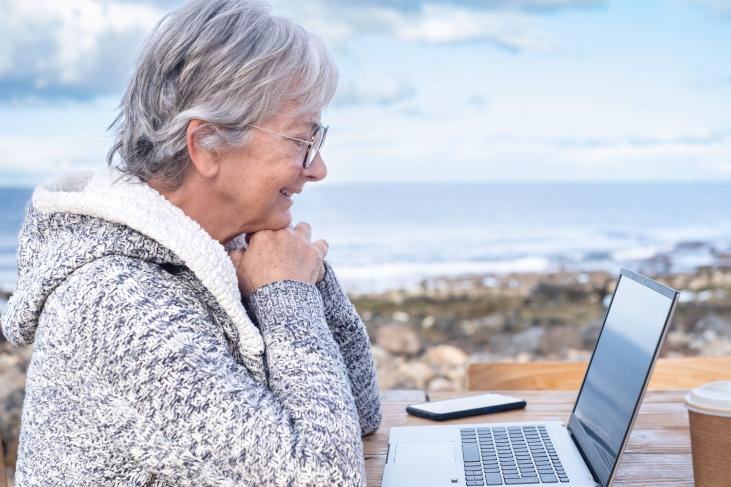 Older woman with gray hair and glasses, wearing a cozy sweater, sits outdoors by the sea, smiling at a laptop on a wooden table. A smartphone and coffee cup are also on the table.
