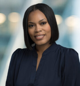 A woman with straight, shoulder-length black hair and a navy blouse smiles softly at the camera against a blurred, modern indoor background with lights.