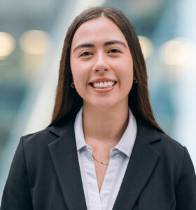 A young woman with long brown hair, wearing a black blazer over a light blue blouse, smiles at the camera. The background is blurred with blue and white tones.