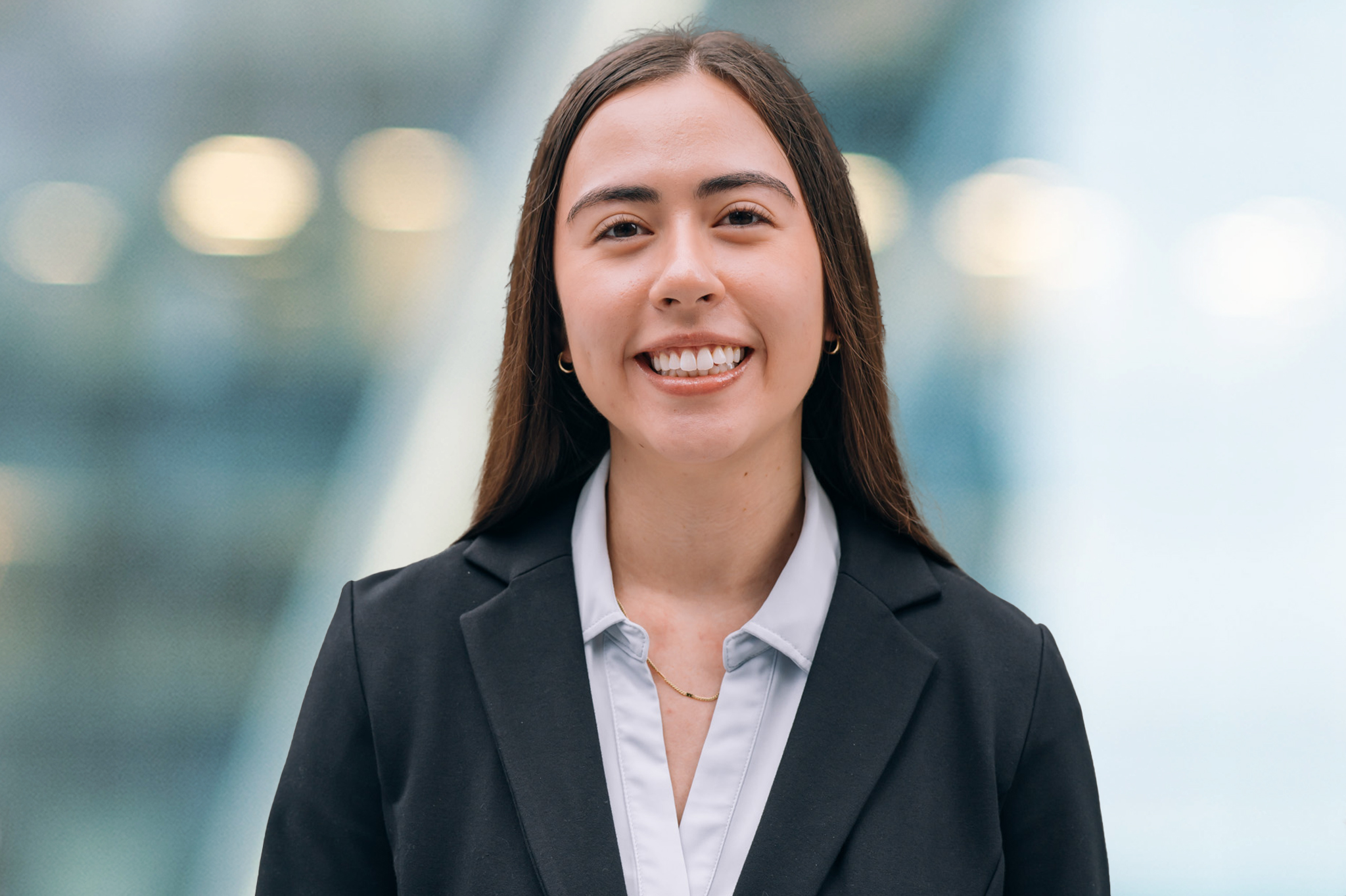 A young woman with long brown hair, wearing a black blazer over a light blue blouse, smiles at the camera. The background is blurred with blue and white tones.