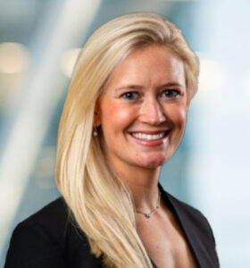 A woman with long blonde hair wearing a black blazer and a necklace smiles in front of a blurred, light-filled background.