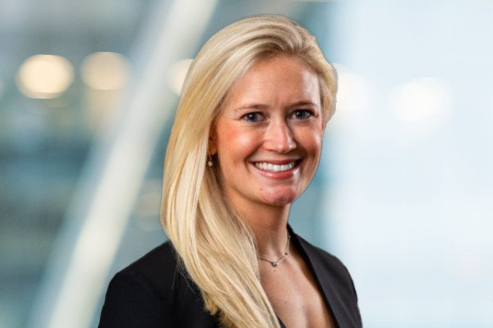 A woman with long blonde hair wearing a black blazer and a necklace smiles in front of a blurred, light-filled background.