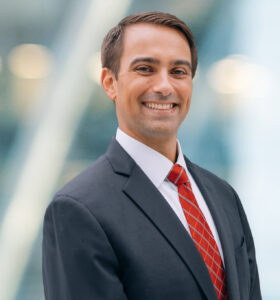 A man in a dark suit, white shirt, and red striped tie smiles while standing in front of a blurred, modern glass building background.