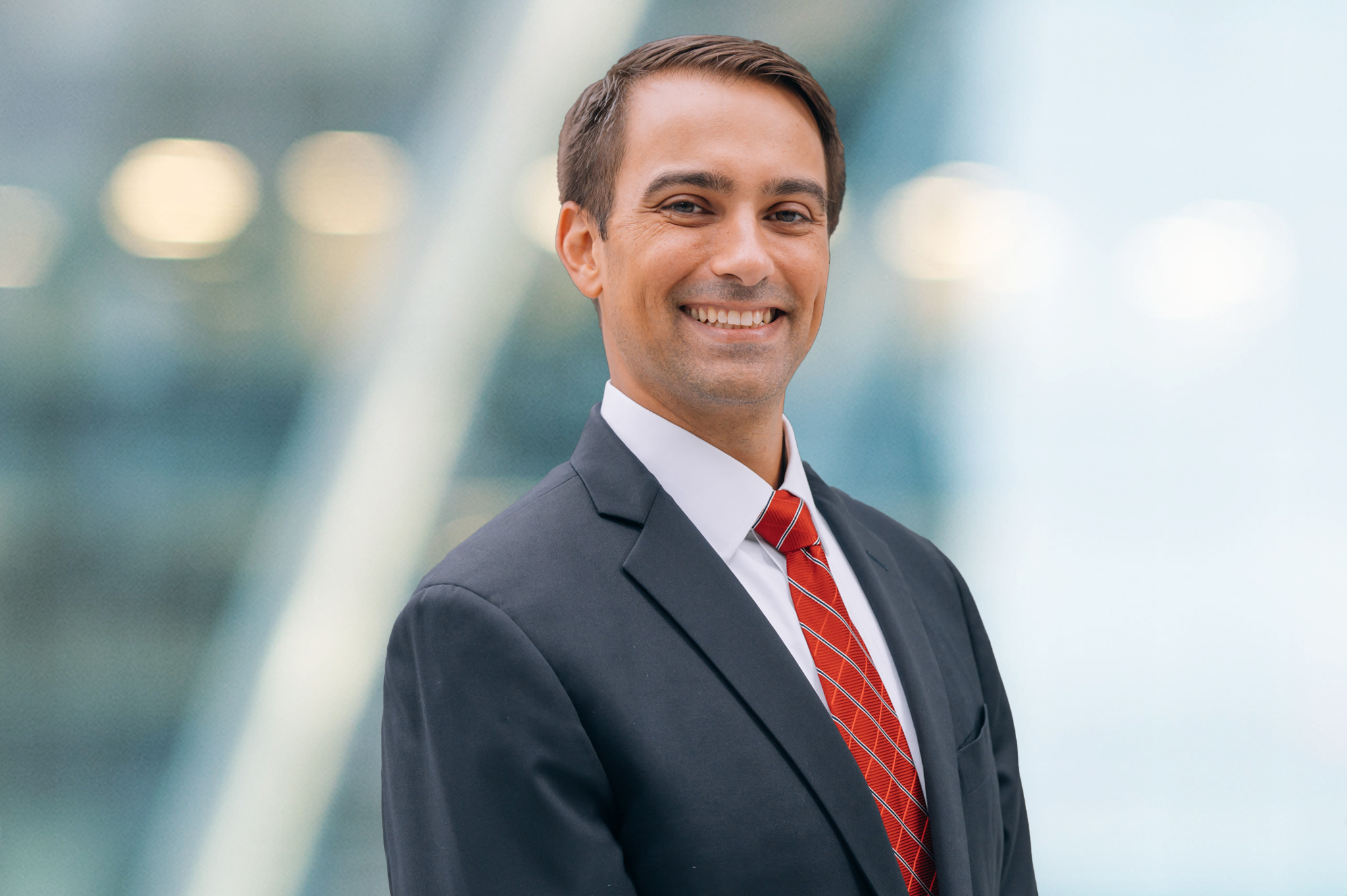 A man in a dark suit, white shirt, and red striped tie smiles while standing in front of a blurred, modern glass building background.