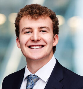 A young man with curly hair wearing a dark suit, white shirt, and patterned tie smiles at the camera against a blurred, modern background with blue and white tones.