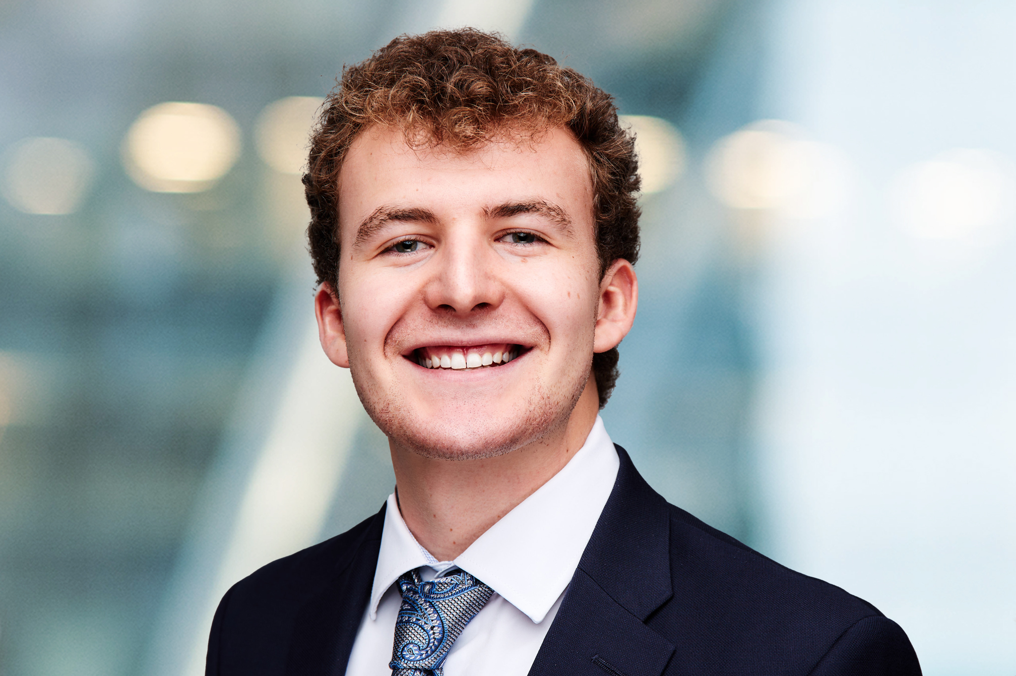 A young man with curly hair wearing a dark suit, white shirt, and patterned tie smiles at the camera against a blurred, modern background with blue and white tones.