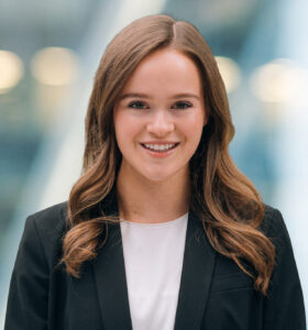 A young woman with long brown hair, wearing a black blazer and white top, smiles at the camera against a blurred modern indoor background with lights.