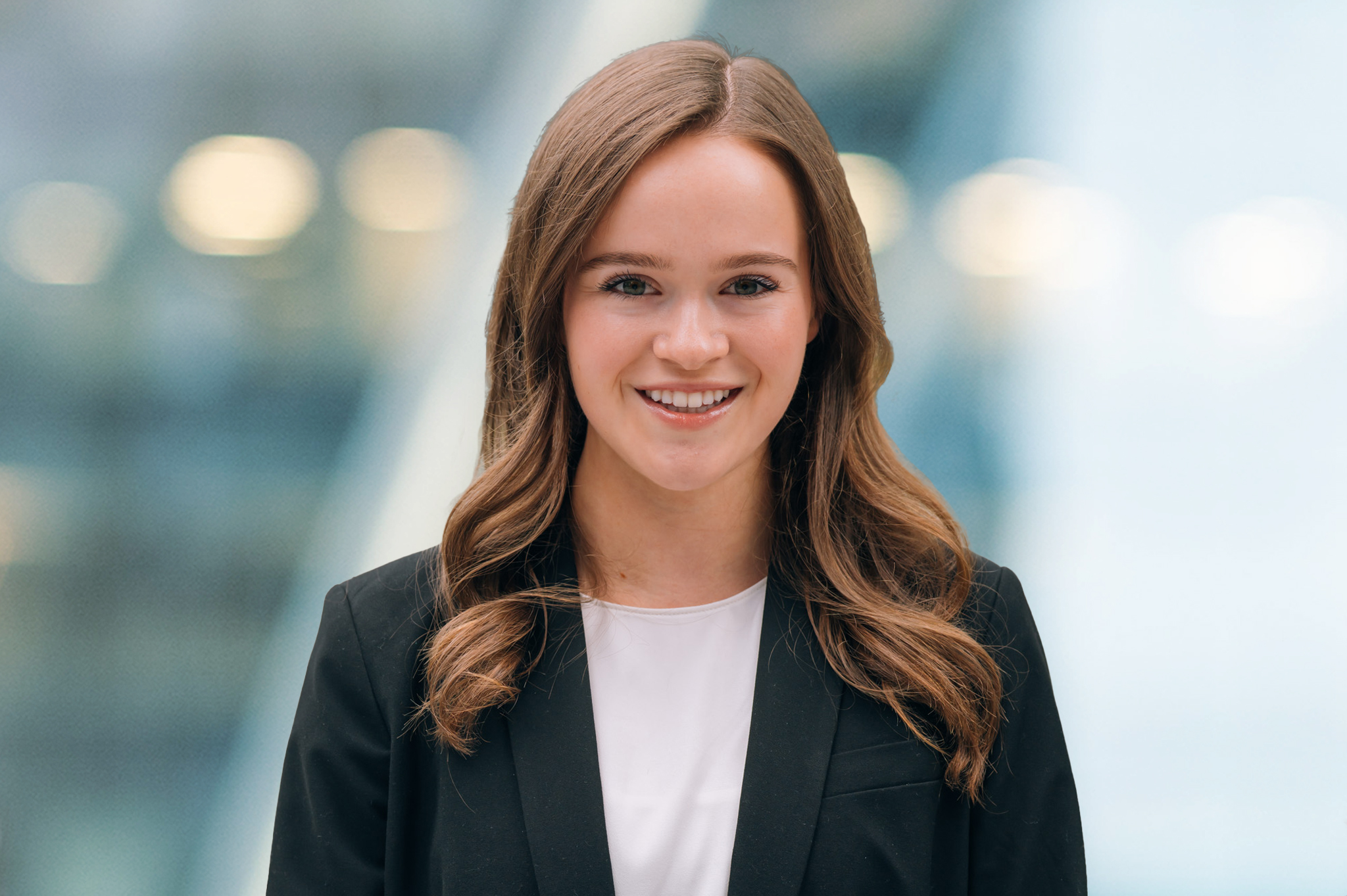 A young woman with long brown hair, wearing a black blazer and white top, smiles at the camera against a blurred modern indoor background with lights.