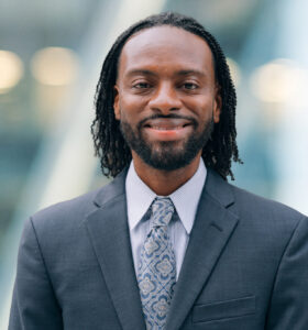 A man with medium-length curly hair and a beard, wearing a dark suit, patterned tie, and light shirt, smiles while standing in front of a blurred modern background.