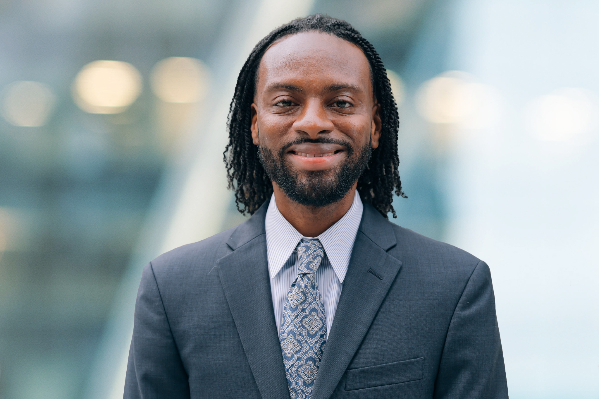 A man with medium-length curly hair and a beard, wearing a dark suit, patterned tie, and light shirt, smiles while standing in front of a blurred modern background.
