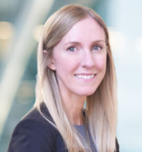 A woman with long blonde hair smiles at the camera. She is wearing a dark blazer over a gray top, and the background is softly blurred with light and glass elements.
