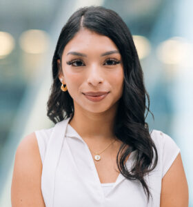 A young woman with long dark hair and light makeup is wearing a white blouse and gold jewelry, standing in front of a blurred modern background with lights. She is looking at the camera and smiling softly.