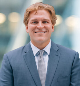 A smiling man in a blue suit and light patterned tie stands in front of a blurred background with soft lights.