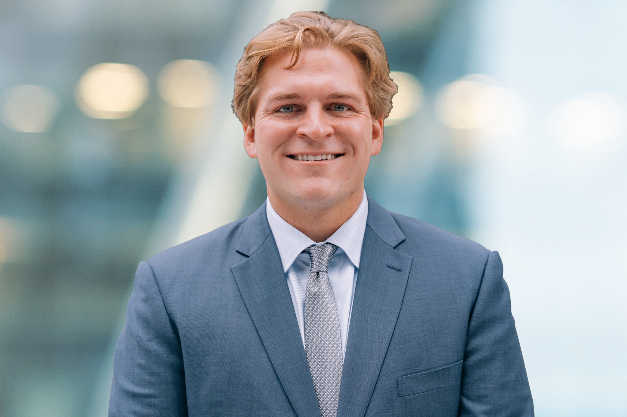 A smiling man in a blue suit and light patterned tie stands in front of a blurred background with soft lights.