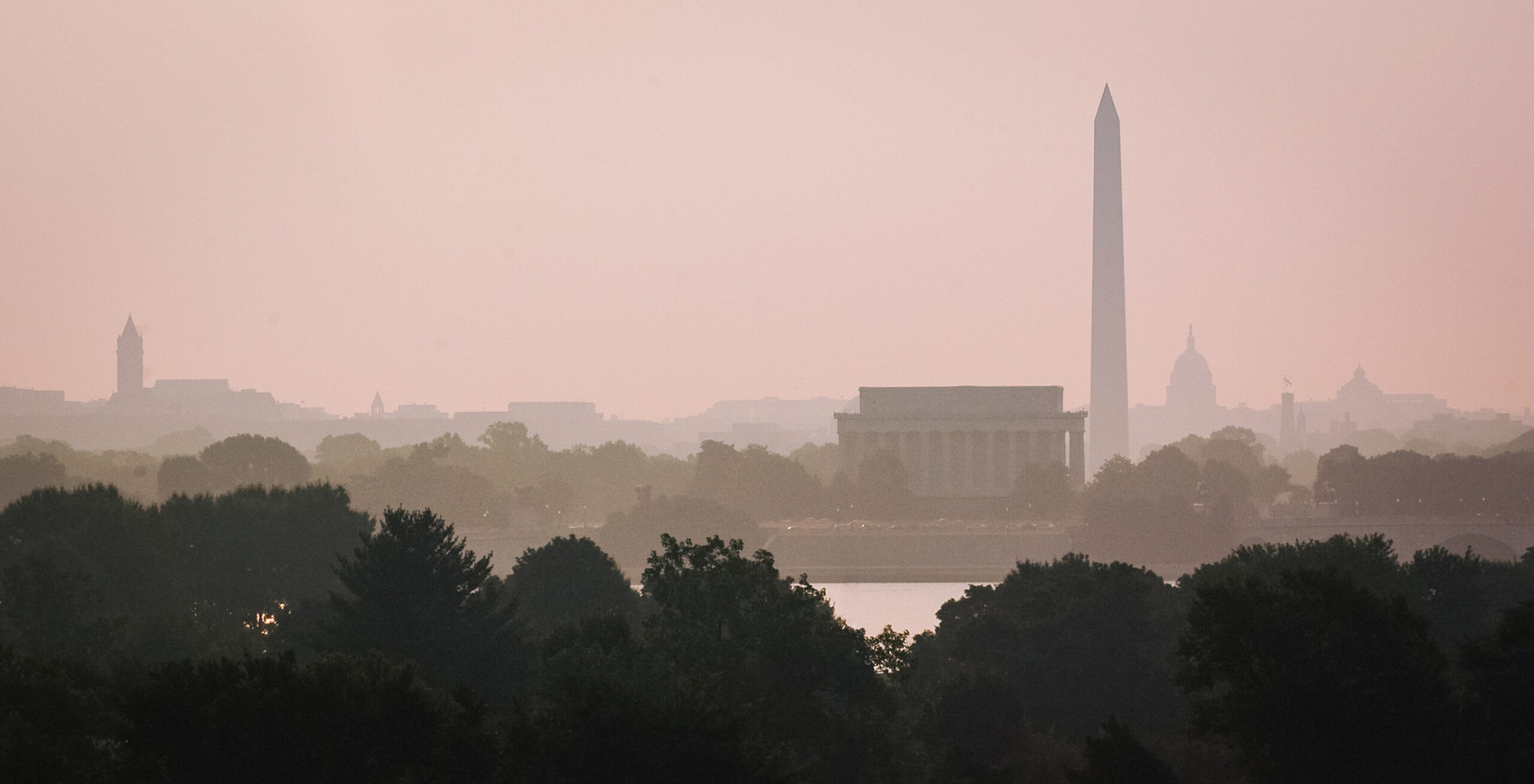 Washington, D.C. skyline at sunrise with the Washington Monument, Lincoln Memorial, and Capitol building shrouded in a pinkish haze, and trees in the foreground.