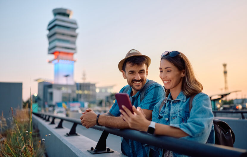 A smiling man and woman stand by a railing outside an airport terminal at sunset, looking at a smartphone together. The man wears a hat and the woman wears sunglasses. An air traffic control tower is in the background.
