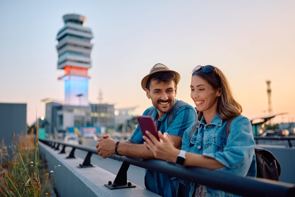 A smiling man and woman stand by a railing outside an airport terminal at sunset, looking at a smartphone together. The man wears a hat and the woman wears sunglasses. An air traffic control tower is in the background.