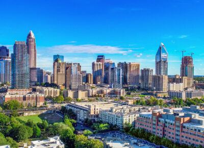 Aerial view of a modern city skyline with tall skyscrapers, mid-rise buildings, green trees, and clear blue sky on a sunny day.
