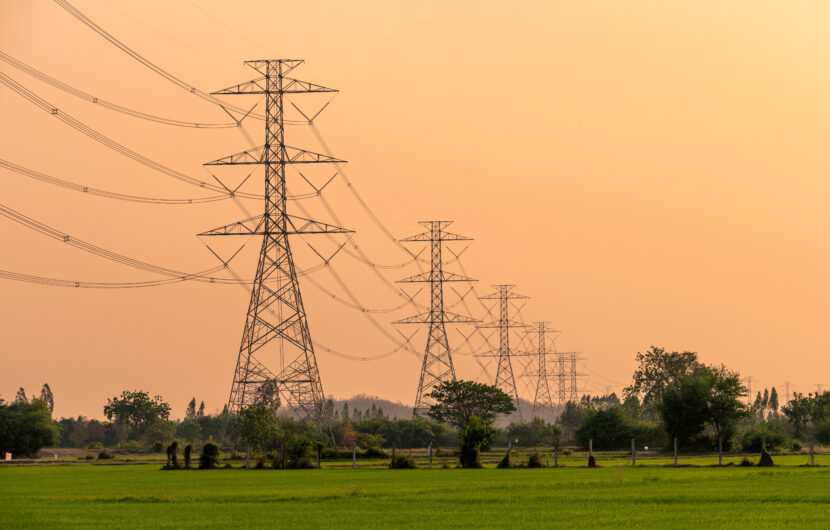 Tall electricity pylons stand in a row across a green field, with power lines stretching into the distance under an orange sky at sunset. Trees and distant vegetation are visible in the background.