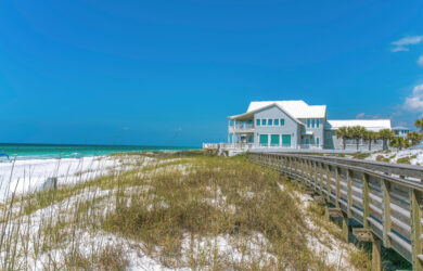 A modern beach house with a white roof sits beside a wooden boardwalk on a sandy beach with grass and turquoise ocean under a clear blue sky.