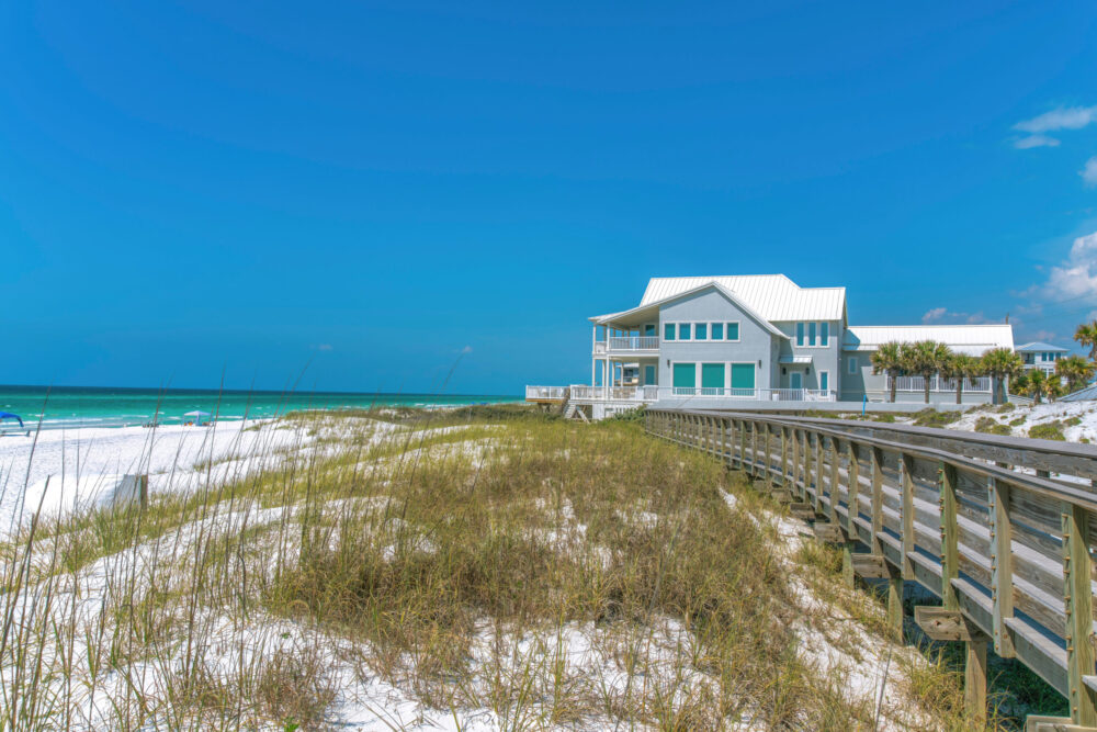 Boardwalk and beach house near the white sand shore of a beach at Destin, Florida A modern beach house with a white roof sits beside a wooden boardwalk on a sandy beach with grass and turquoise ocean under a clear blue sky.