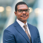 A man wearing glasses, a blue suit, a white shirt, and a striped tie smiles slightly while standing against a blurred background with soft lights.
